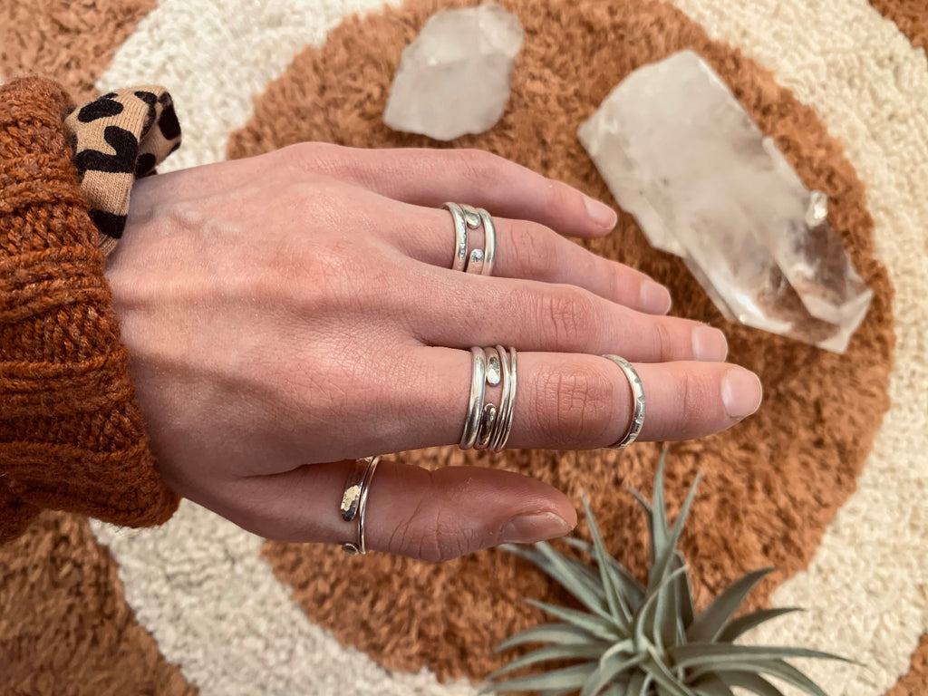 Image shows a hand wearing lots of textured silver stacking rings. The hand reaches over a rusty brown rug with a white geometric design. Quartz crystals and air plants accent the image.  