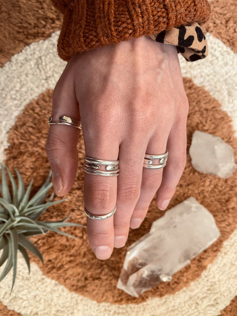 A hand reaches into the frame from the top. She is layering many simple, textured silver rings on multiple fingers. In the background are crystals and air plants.