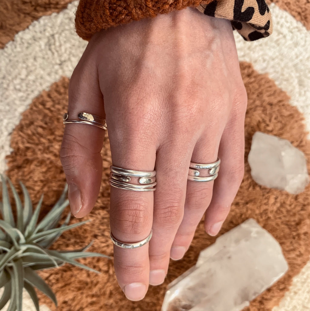 A hand reaches into the frame from the top of the image. The hand is decked out in sterling silver stacking bands, layered in unique stacking ring combinations. The rings are in different widths, with hammered and filed details. The background of the image is a Morrow Soft Goods bathmat in brown and cream. Clear quartz crystals and air plants accent the image. The cuff of a rust orange knit sweater, and a leopard print arq scrunchie are peeking into the shot.