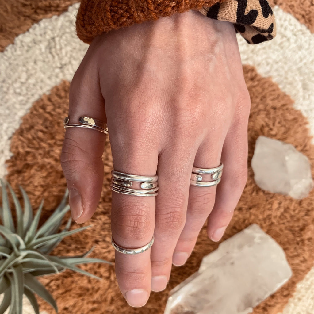 Hand wearing various silver stacking ring bands. Brown  circular fuzzy backdrop with clear crystals and green air plants.