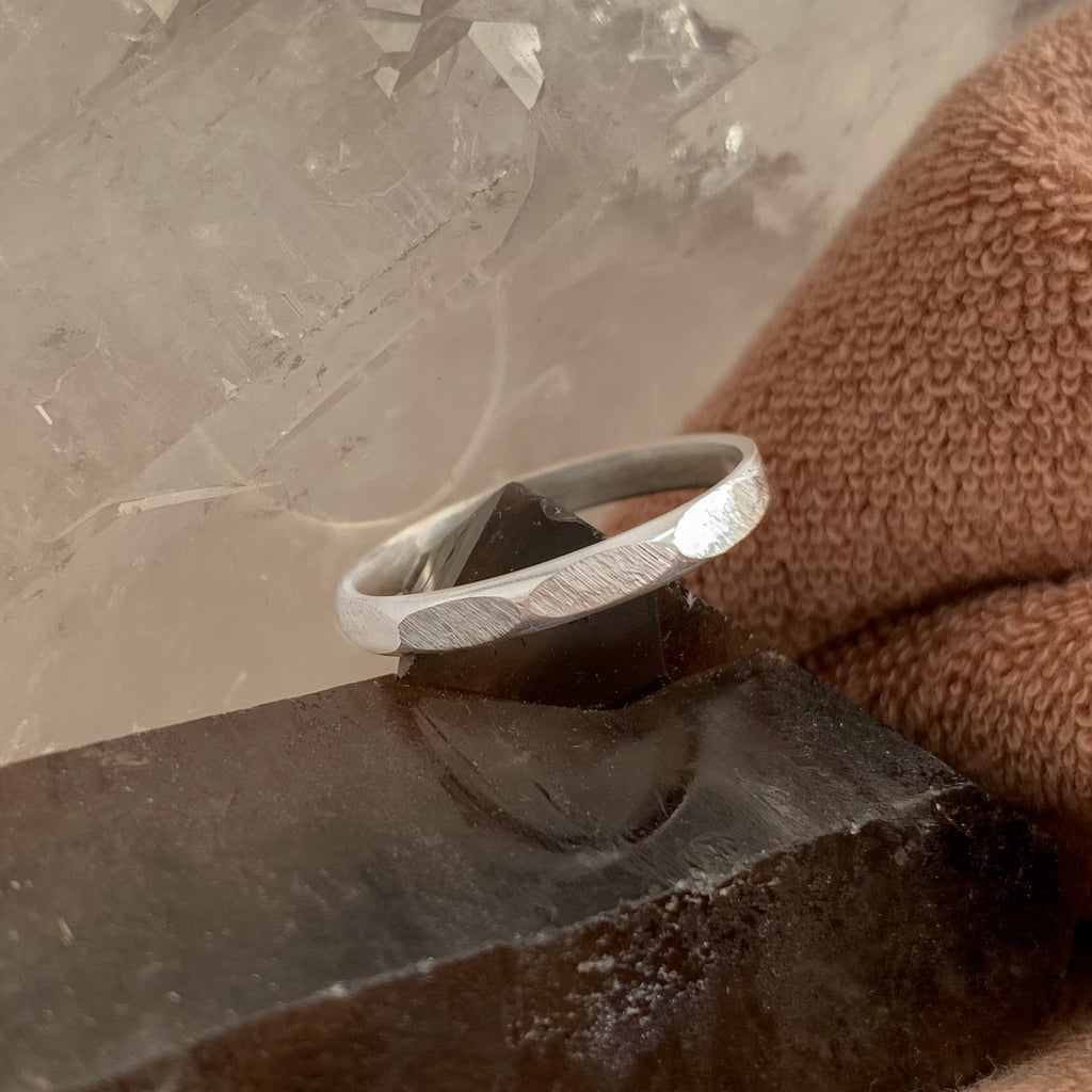 Geometric stacking ring with hand-filed facets. The traditional metalsmithing tools show in the file marks on the handmade silver band. The half-round ring band balances on a dark smoky quartz crystal. Brown terry cloth in the right of the image, and clear quartz towards the top left.