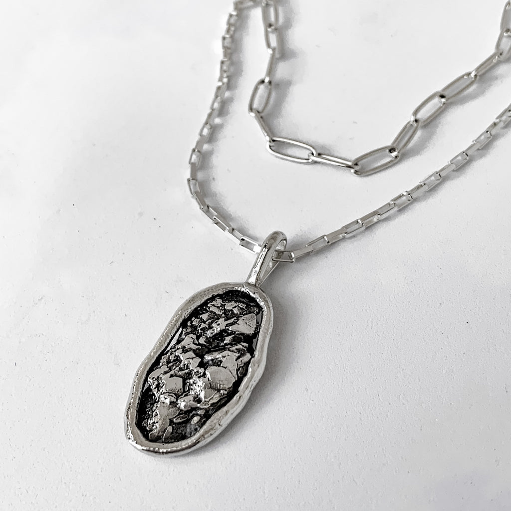 Oval-shaped recycled silver charm on a layered silver chain. Geology inspired pendant has a smooth raised border and is on a silver box chain. The shorter chain layered with it is a large-link oval chain.