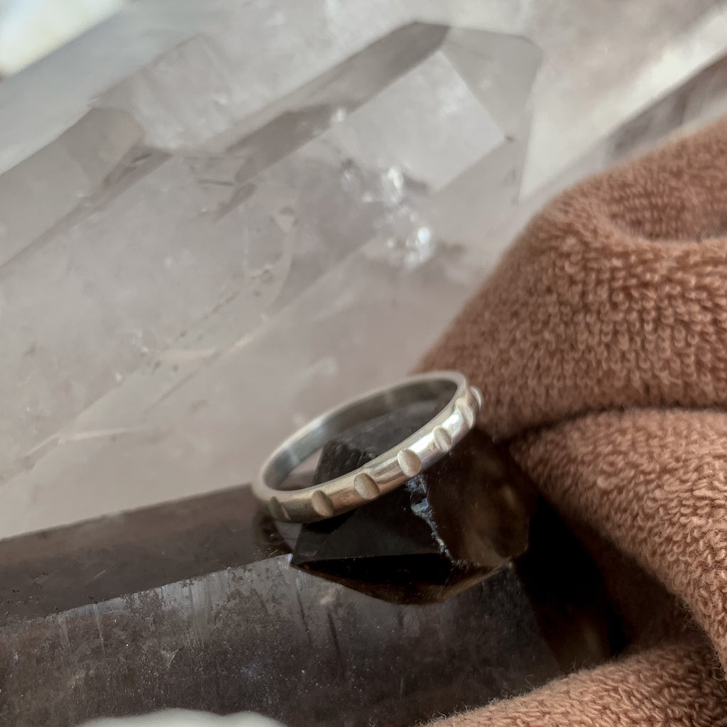 A silver stacking ring with hand-filed dots around the half-round band. The silver ring is posed on a dark smoky quartz crystal, with brown terry fabric and more mineral shapes in the background. The neutral tones complement the cool silver metal.