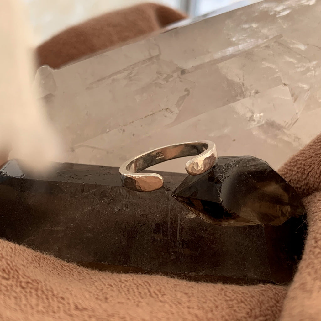 Hammered stacking ring with a gap and rounded edges. A simple ring that layers with other bands or mineral jewelry. The silver band is on a dark grey crystal with brown terry fabric in the front of the shot. The silver reflects the warm neutral tones.
