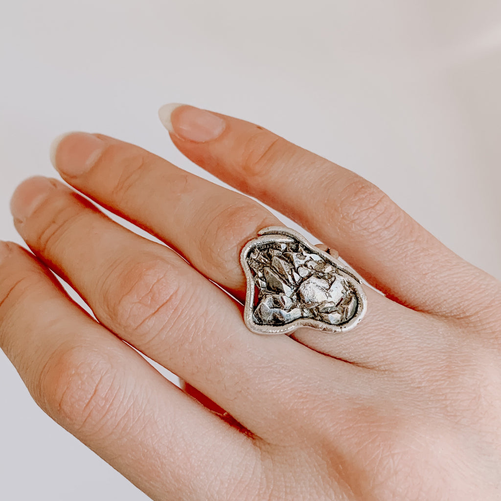 Solid recycled silver ring in an organic design. Shown on a hand with a white background. The ring is an asymmetrical wavy shape with crystal textures inside a silver border.